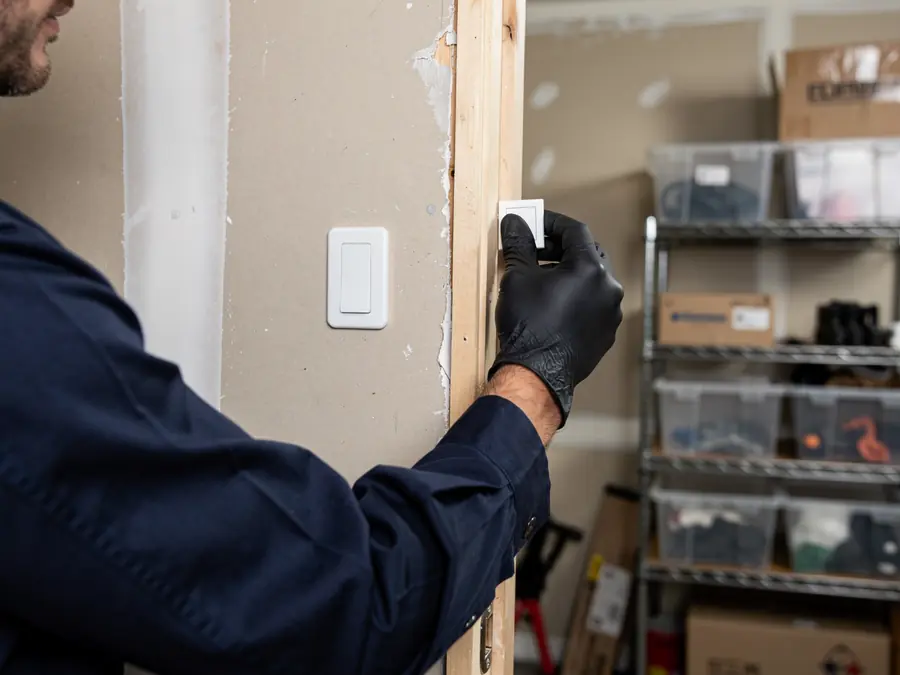 A technician in a blue shirt and black gloves installs a white light switch.